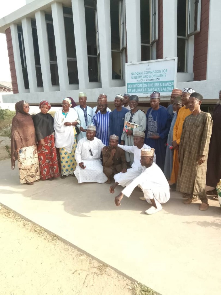 Hon. Aminu Musa with women from the visit, in front of the exhibition sign.